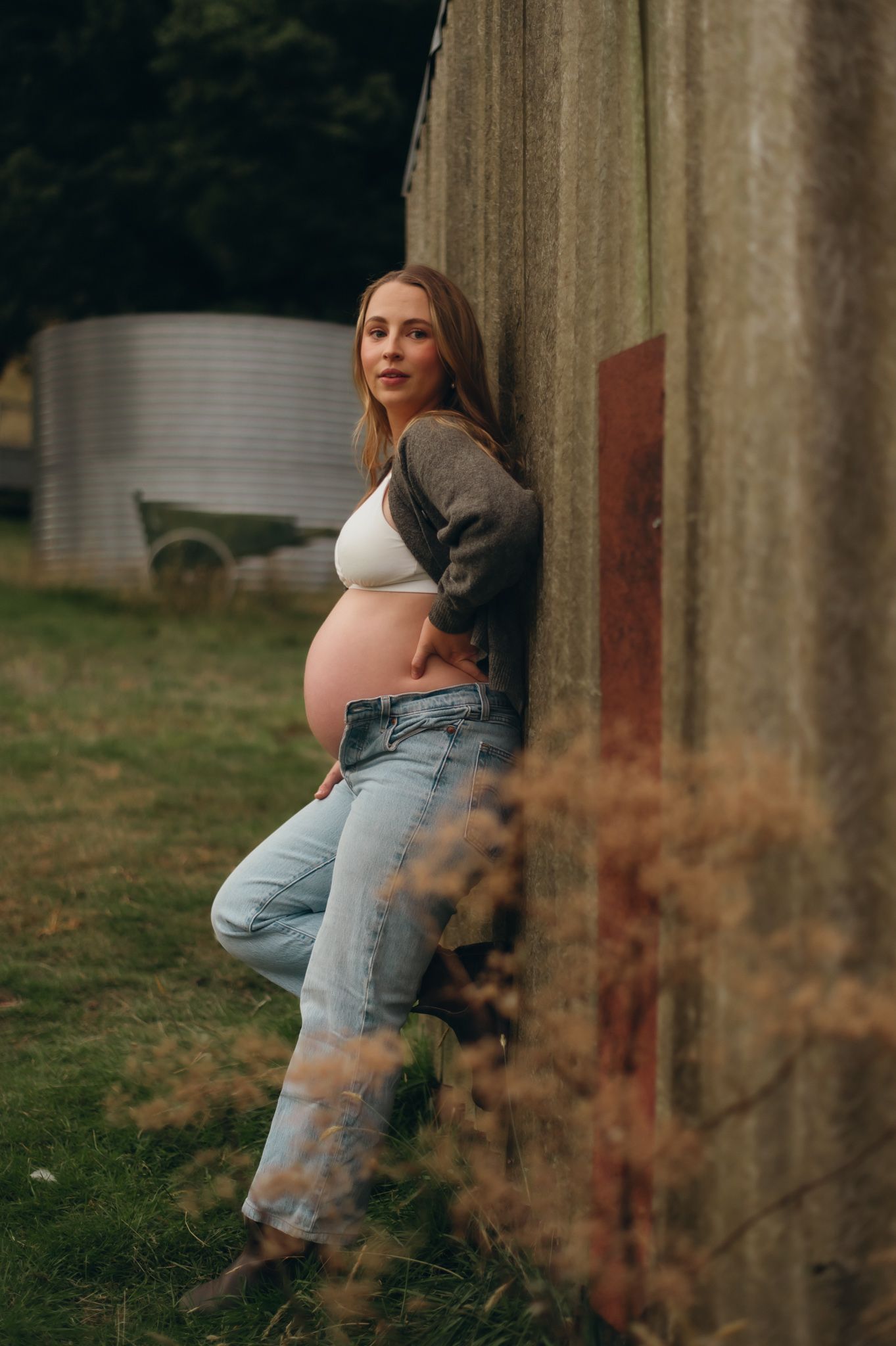 Pregnant woman in jeans and crop top leaning against a rustic barn wall outdoors.