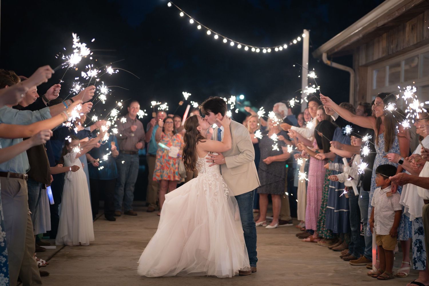 Bride and groom share romantic kiss during sparkler exit at outdoor wedding reception at night.