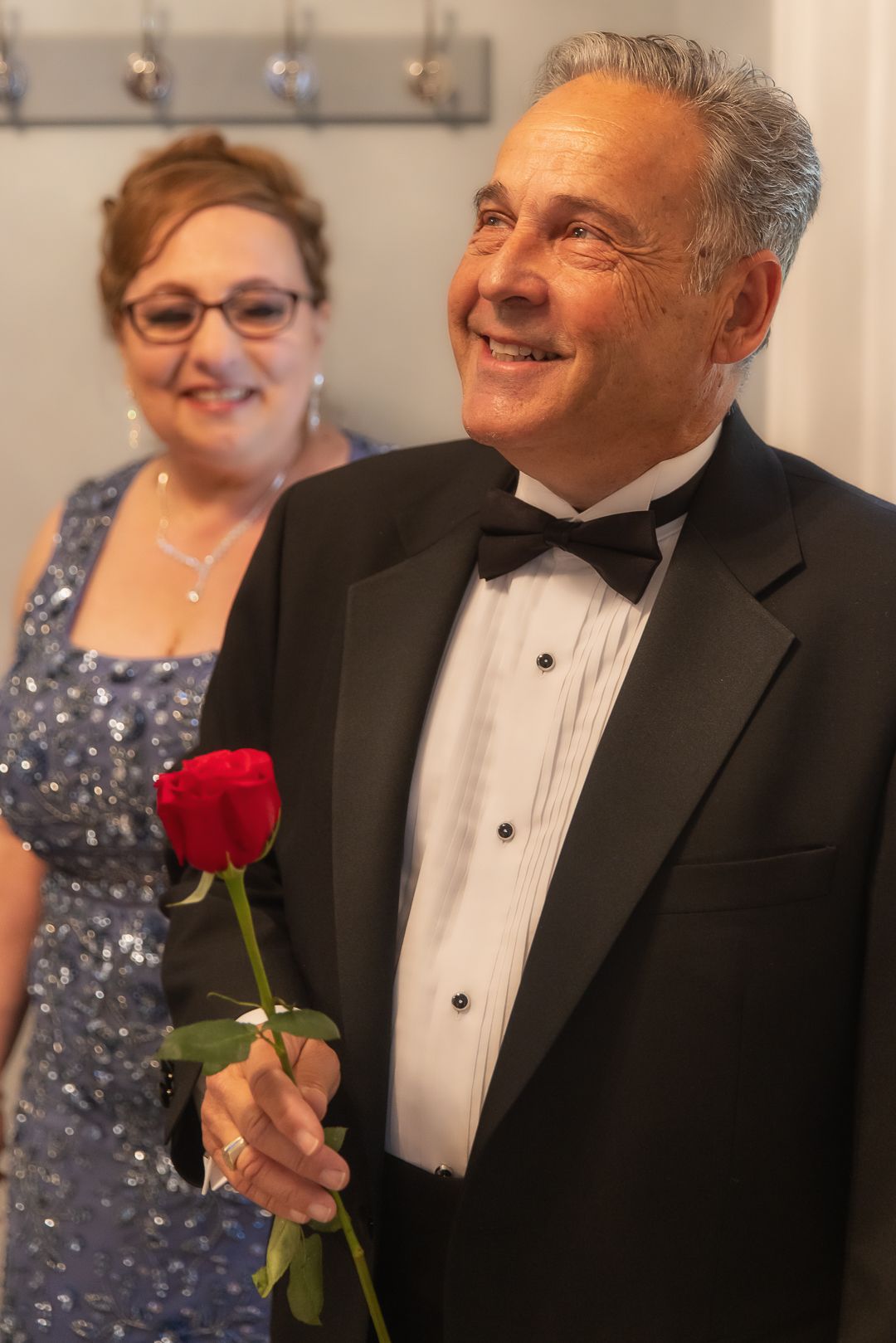 Elegant man in tuxedo holding red rose smiles, woman in blue sequin dress stands behind him.