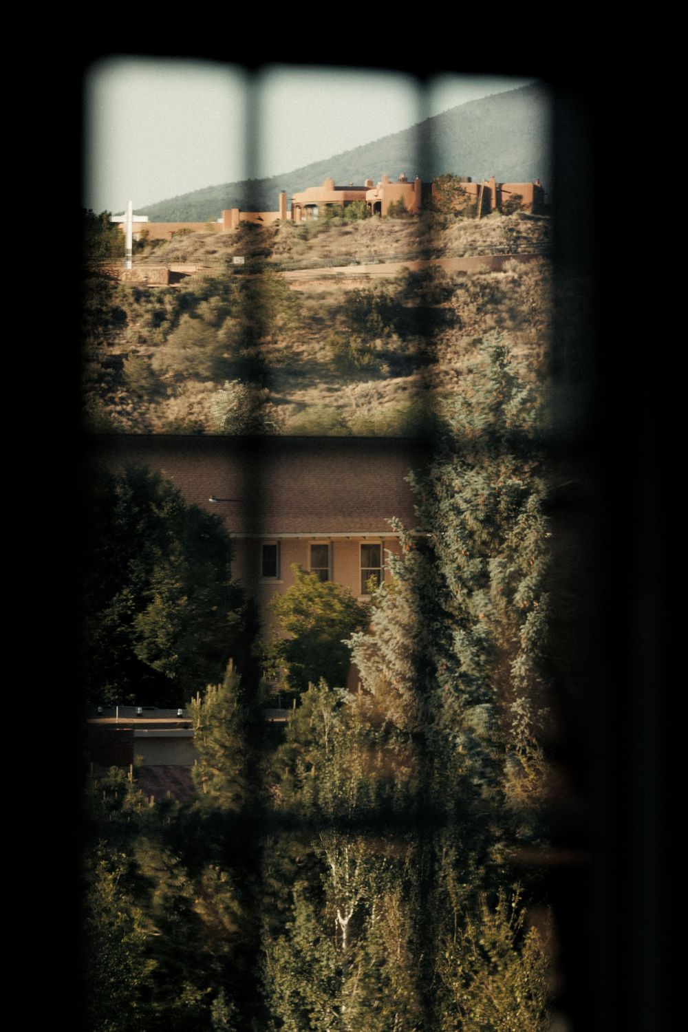View through a barred window overlooking lush trees, a adobe building, and a hillside with a cross in the distance.