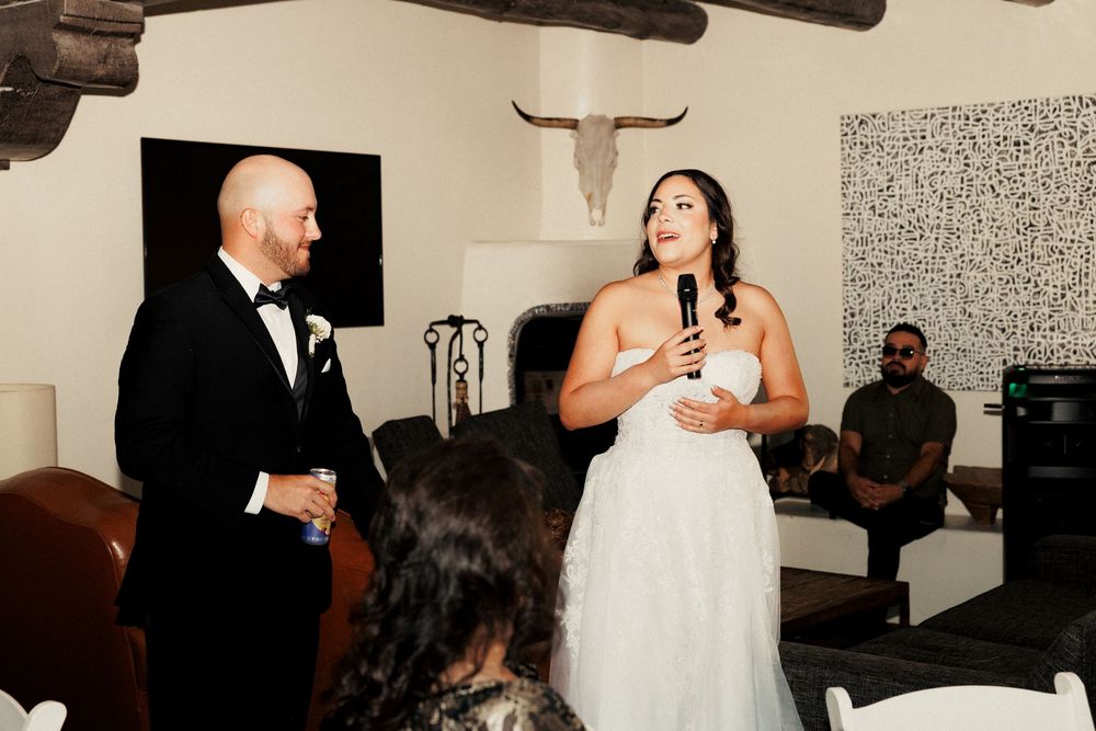 Bride in white gown speaks into microphone beside groom in tuxedo during indoor wedding reception.