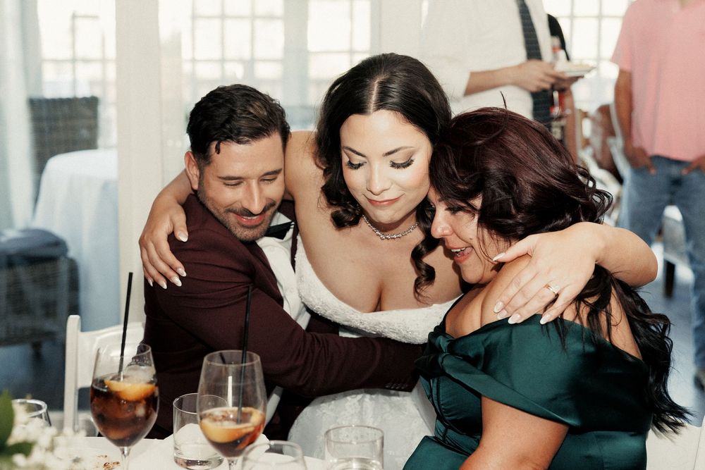 Bride and groom share a warm group hug with a guest in emerald dress at elegant wedding reception.