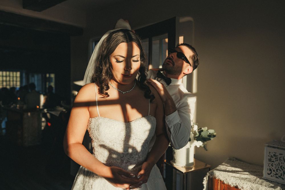 Bride in white strapless gown and veil stands indoors as groom adjusts her dress in warm golden light.