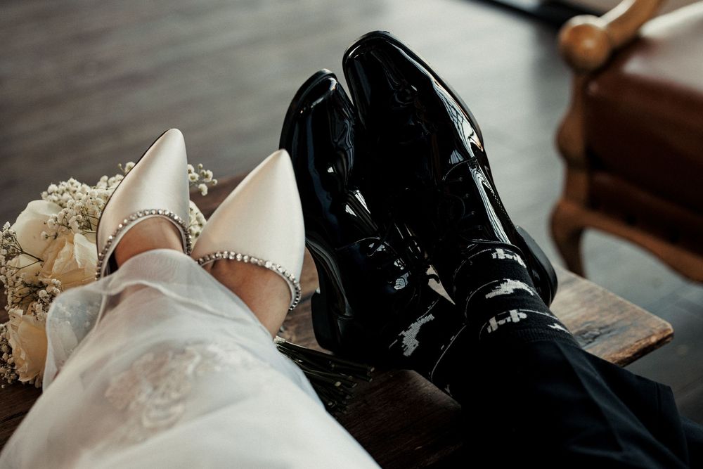 Close-up of bride and groom shoes, white heels with rhinestones and black patent leather dress shoes at a wedding.