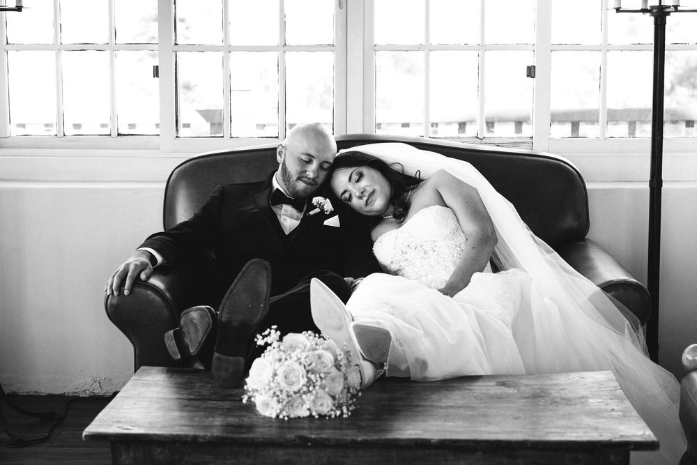 Black and white photo of a bride and groom relaxing on a leather sofa, with a bouquet on the table.