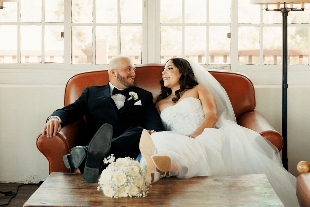 Bride and groom sitting together on a brown leather sofa, smiling at each other, with a white bouquet on the table.