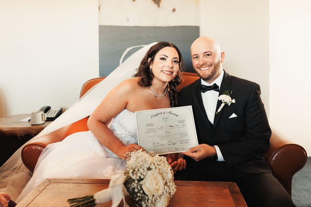 Smiling bride and groom in formal attire holding their marriage certificate while seated on a couch.