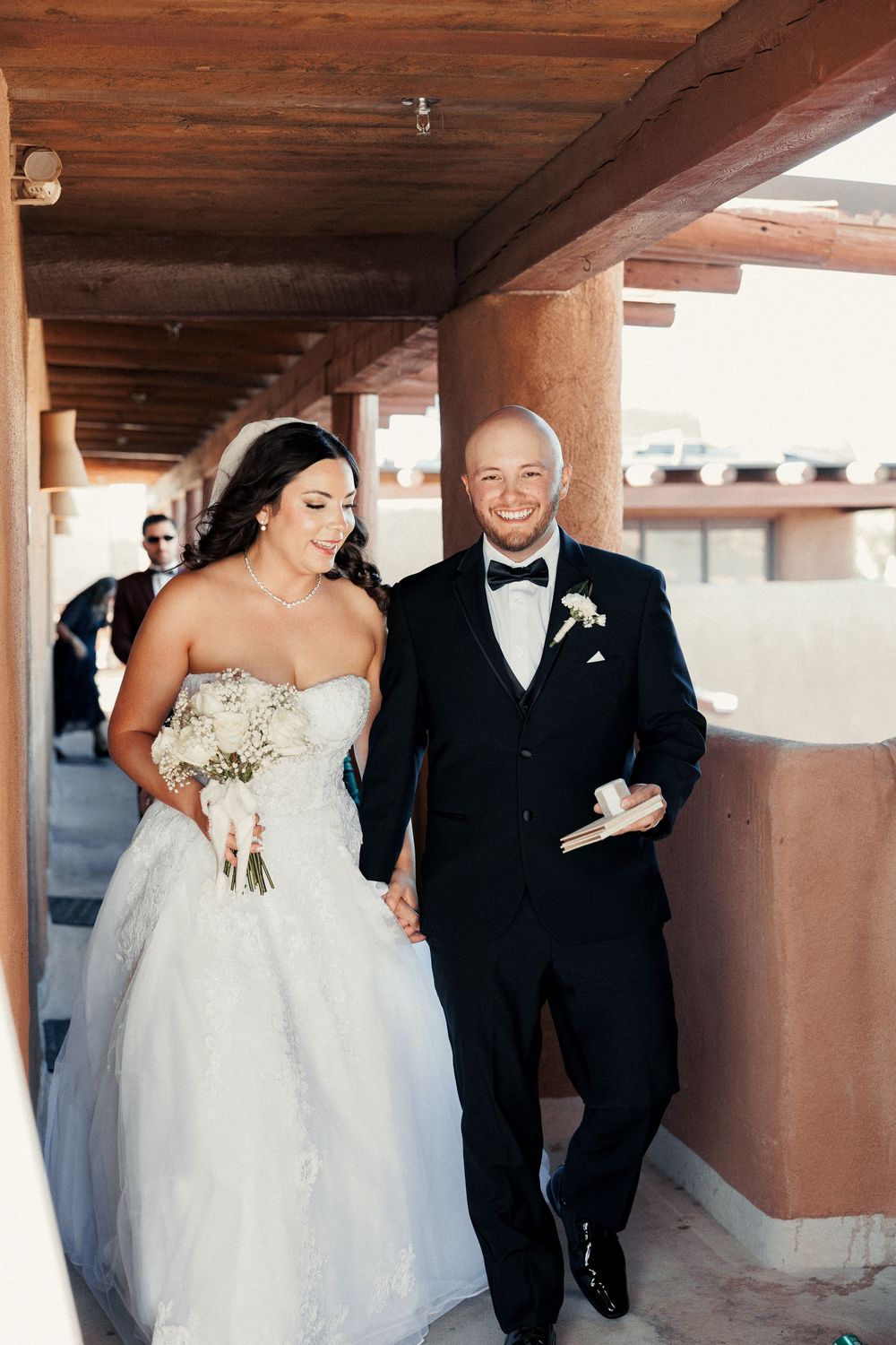 Smiling bride and groom walk together in a covered outdoor corridor, bride holding white bouquet, groom in black tuxedo.