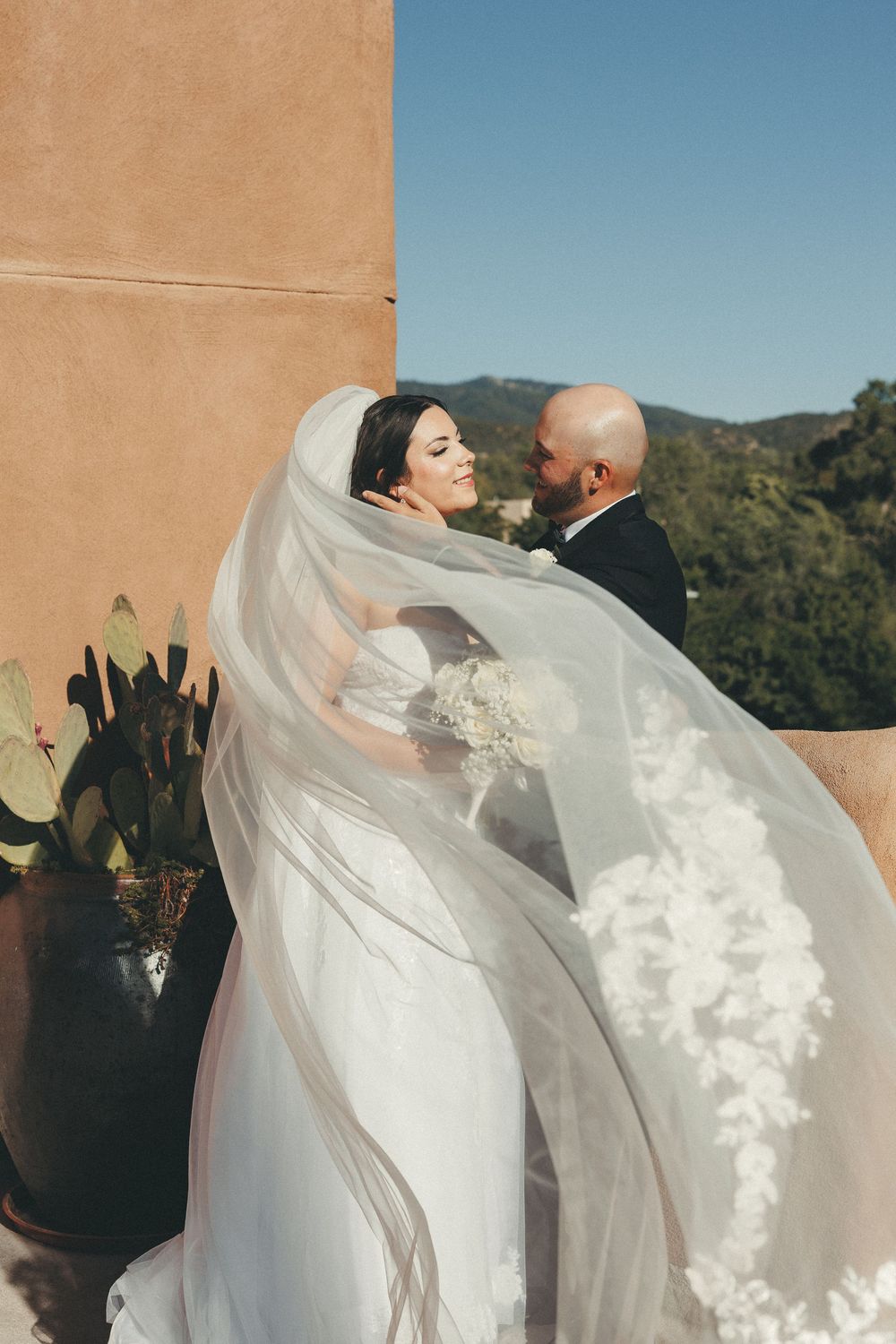Bride in lace wedding dress with flowing veil embraces groom in black tuxedo outdoors with mountain views.