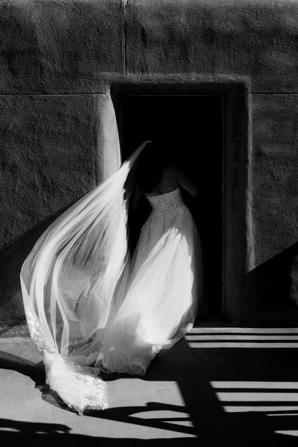 Black and white photo of a bride in a flowing gown and veil standing in a dramatic doorway with light and shadow.
