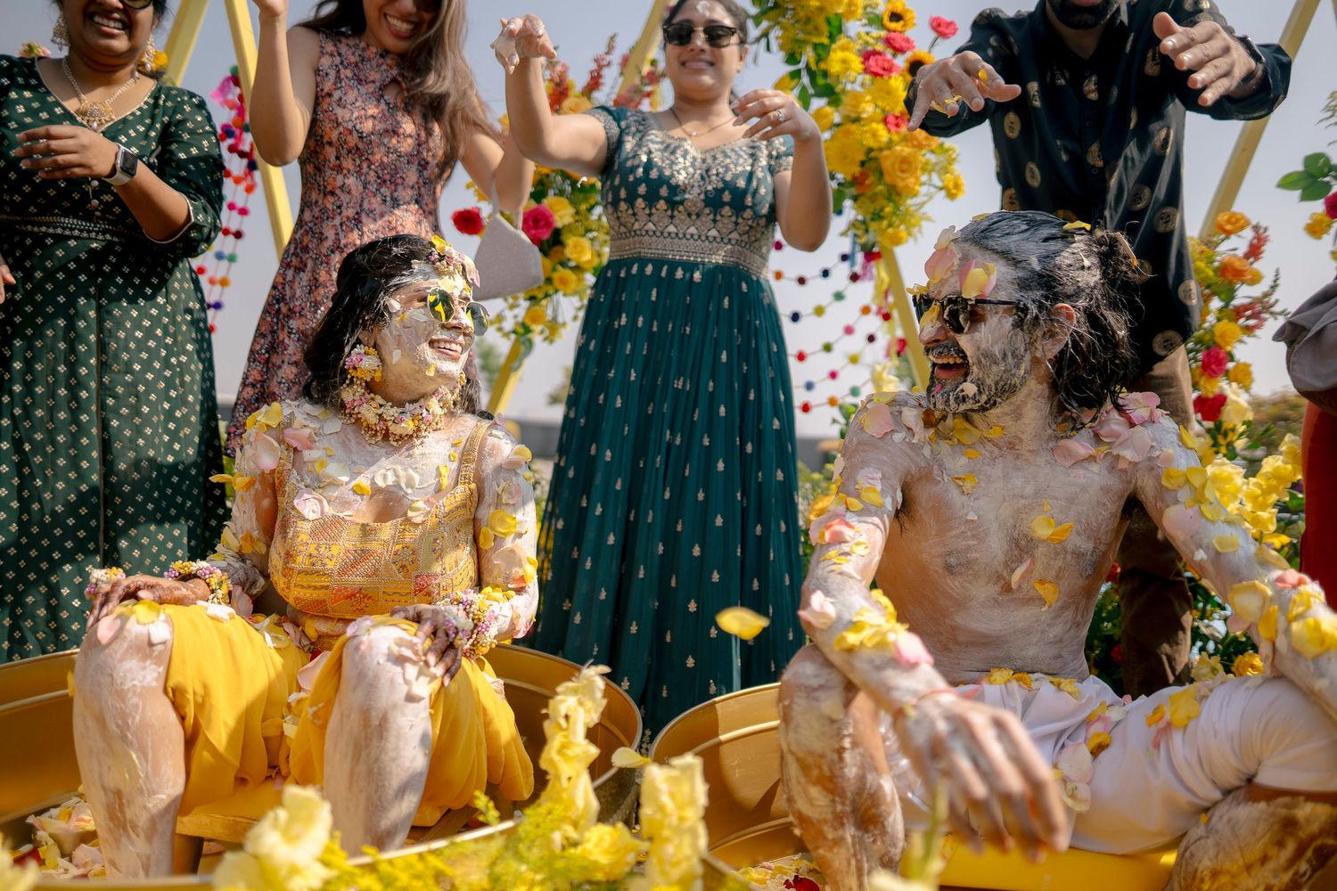 Couple in yellow outfits covered in marigold flowers during Indian Haldi wedding ceremony.