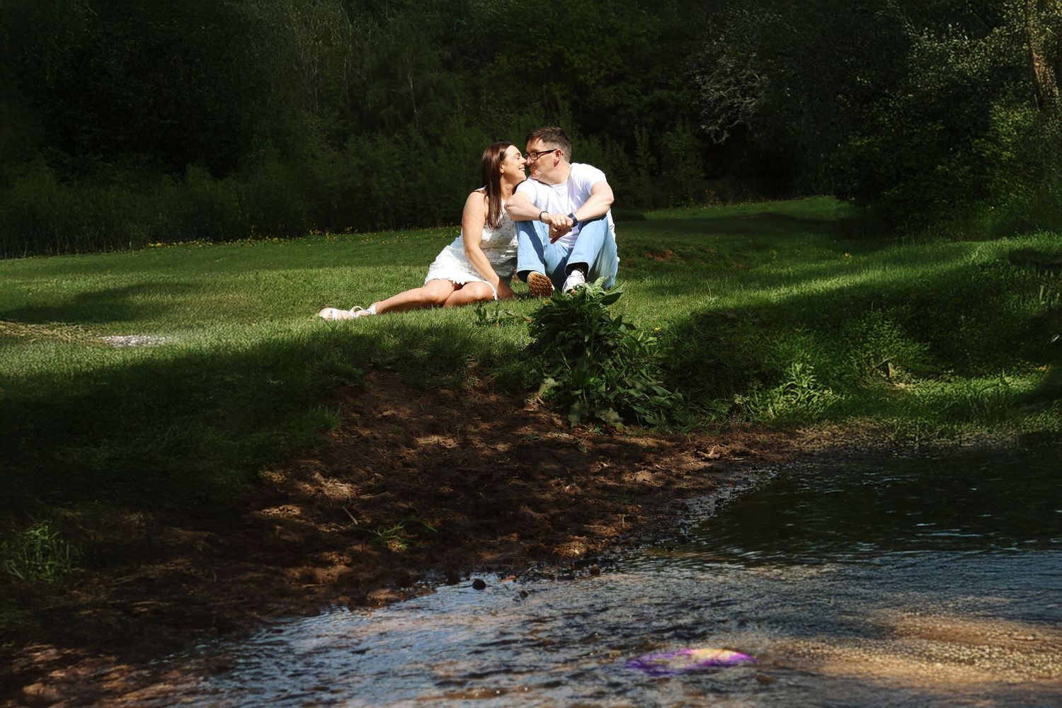 Couple sitting together on grassy riverbank surrounded by lush green trees in a peaceful outdoor setting.