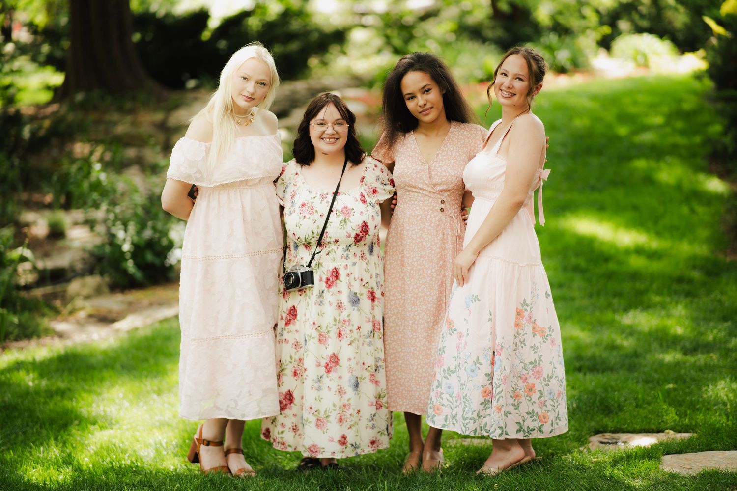 Four women in floral and pastel dresses posing together on green lawn in sunny garden.