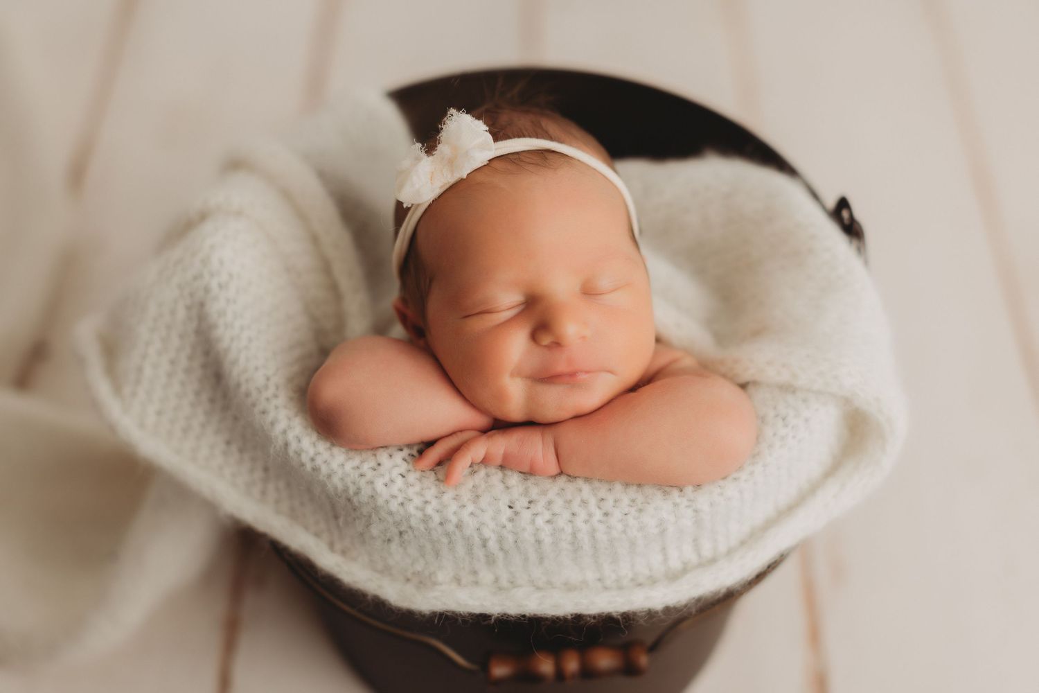 Sleeping newborn baby in a bucket wrapped in a white knit blanket wearing a white bow headband.