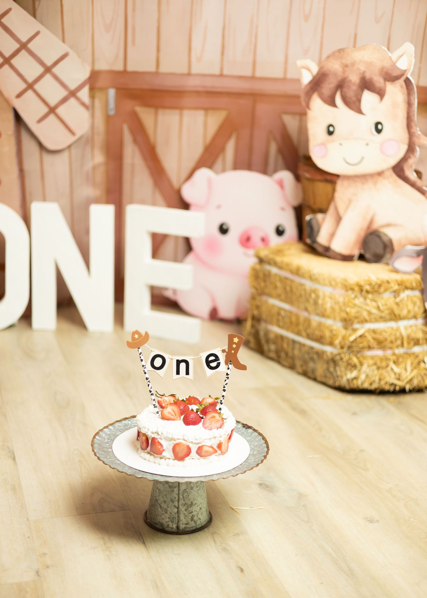 Farm-themed first birthday cake smash setup with stuffed animals, hay bale, and ONE letters on a rustic wooden backdrop.