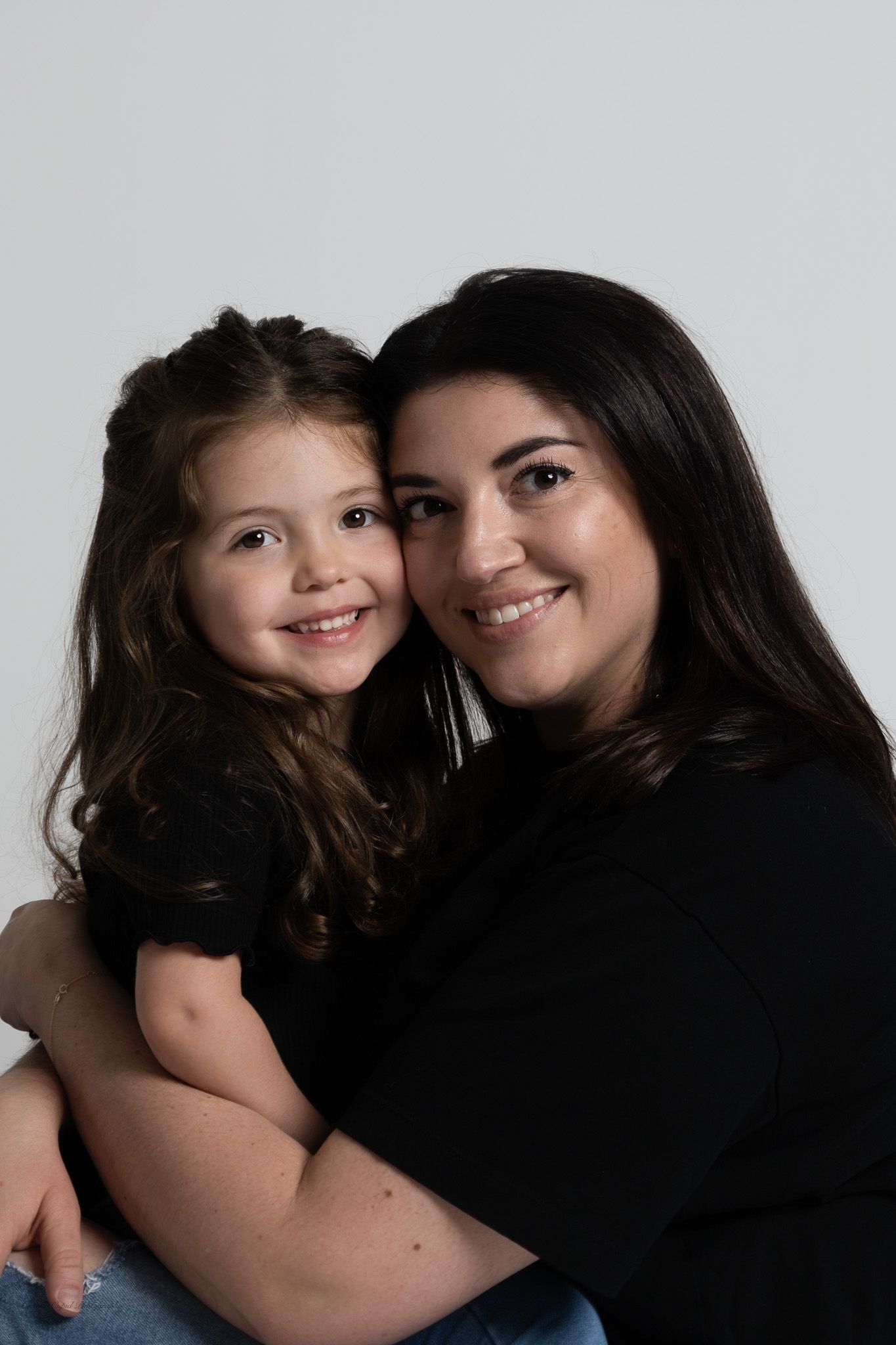 A smiling woman with dark hair hugs a young girl in matching black tops against a white background family portrait session in Pittsburgh