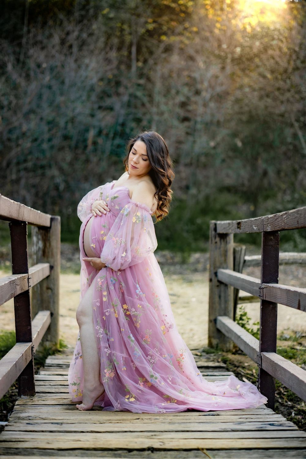 Pregnant woman in flowing pink floral tulle gown posing on a wooden bridge at golden hour.