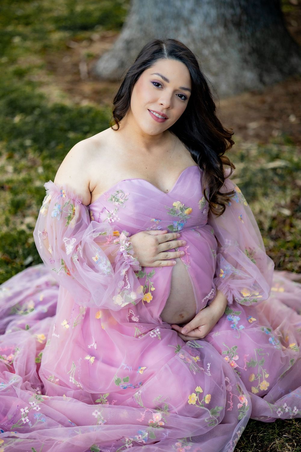 Smiling pregnant woman in pink floral off-shoulder gown sitting outdoors in a lush park setting.