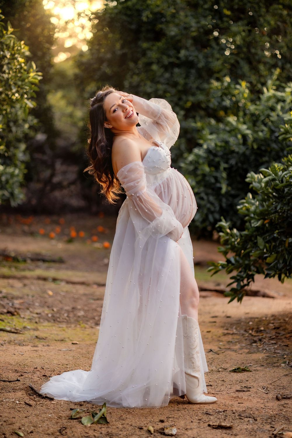 Glowing pregnant woman in white sheer gown smiling in an orange grove at golden hour.