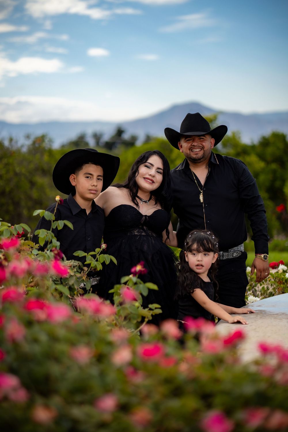 Family of four dressed in all-black western attire with cowboy hats poses among pink roses outdoors.