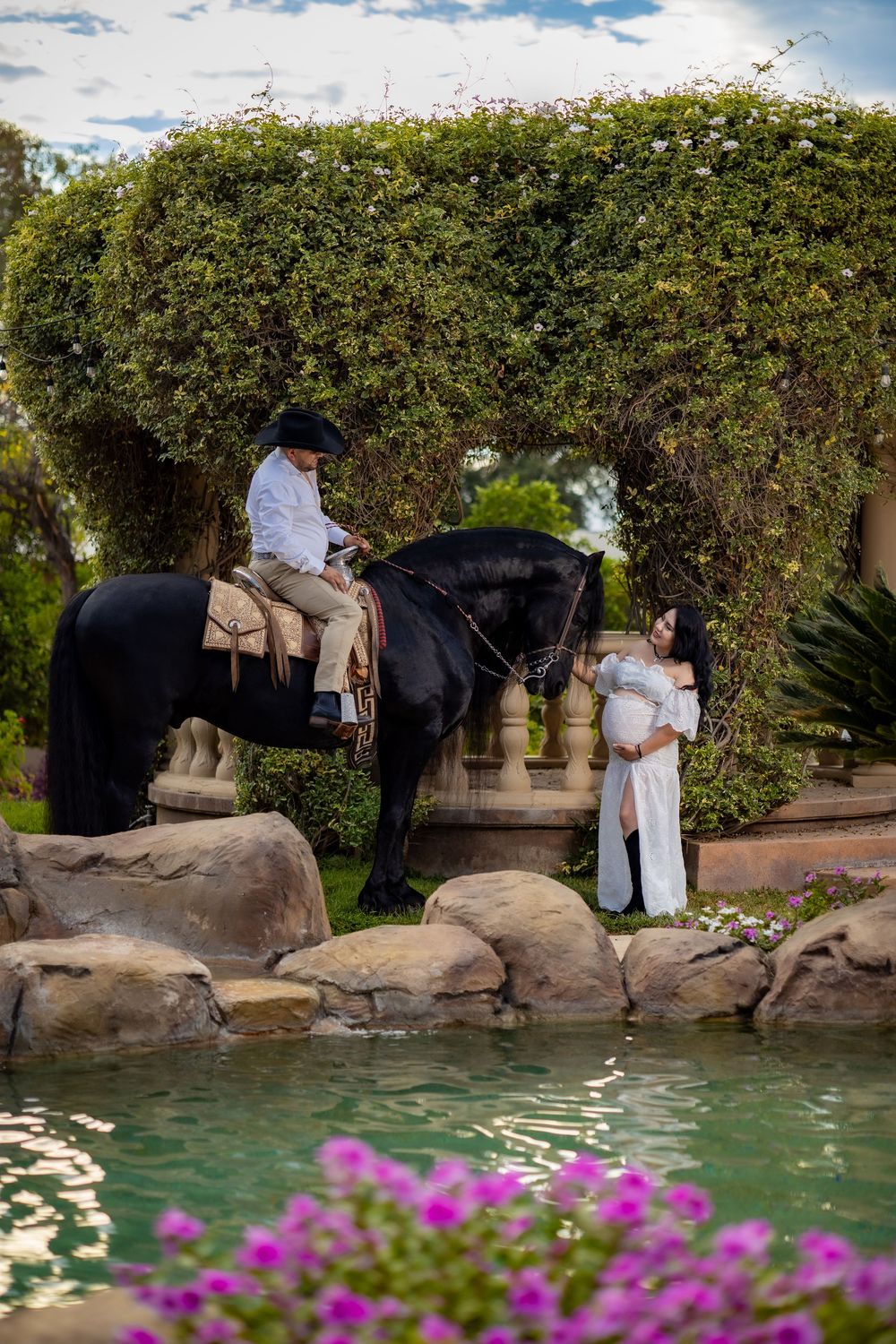 Pregnant woman in white dress stands by pond as man on black horse poses in lush garden.