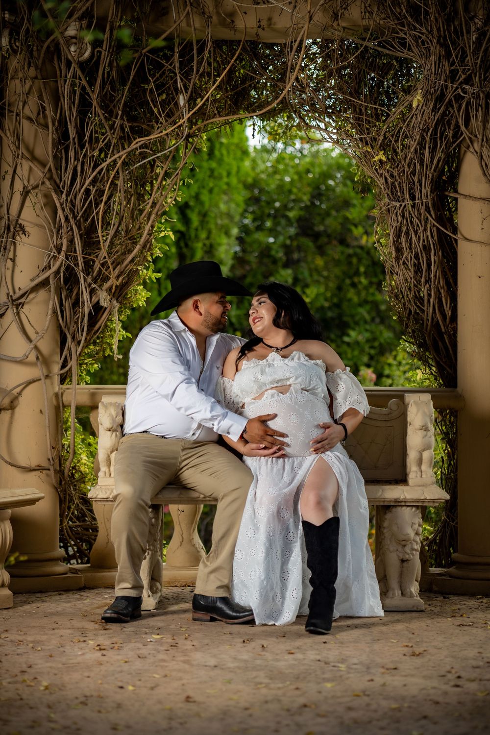 Couple in cowboy hats share tender maternity moment on stone bench under vine-covered garden archway.