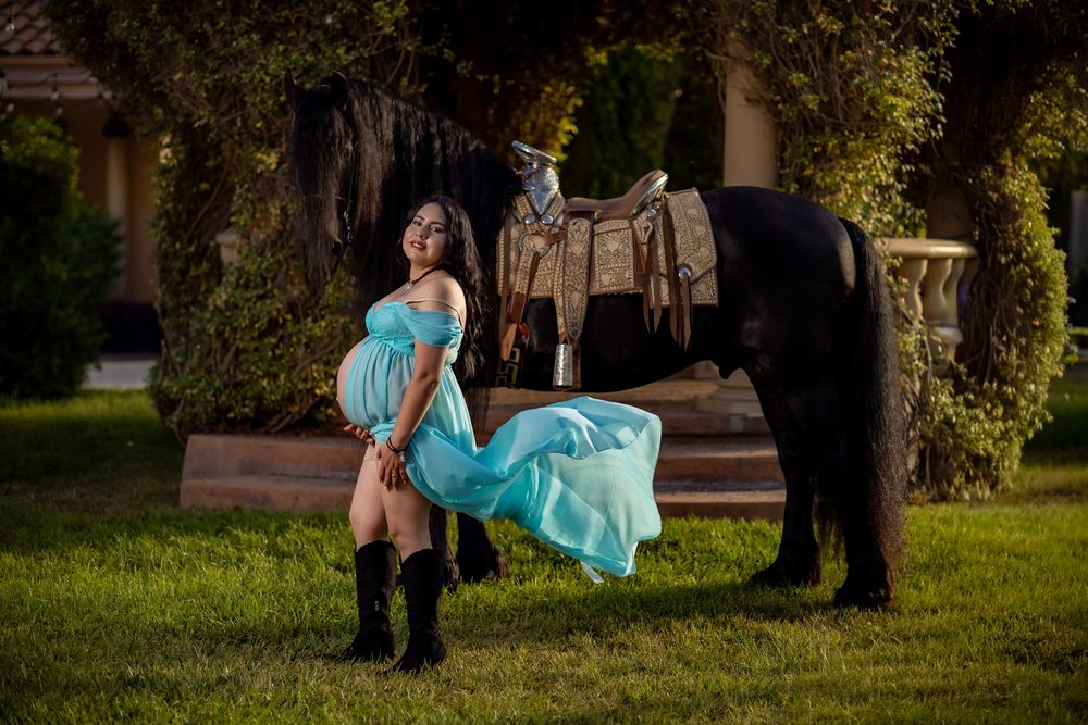 Pregnant woman in teal gown and cowboy boots poses beside a black horse on green grass.