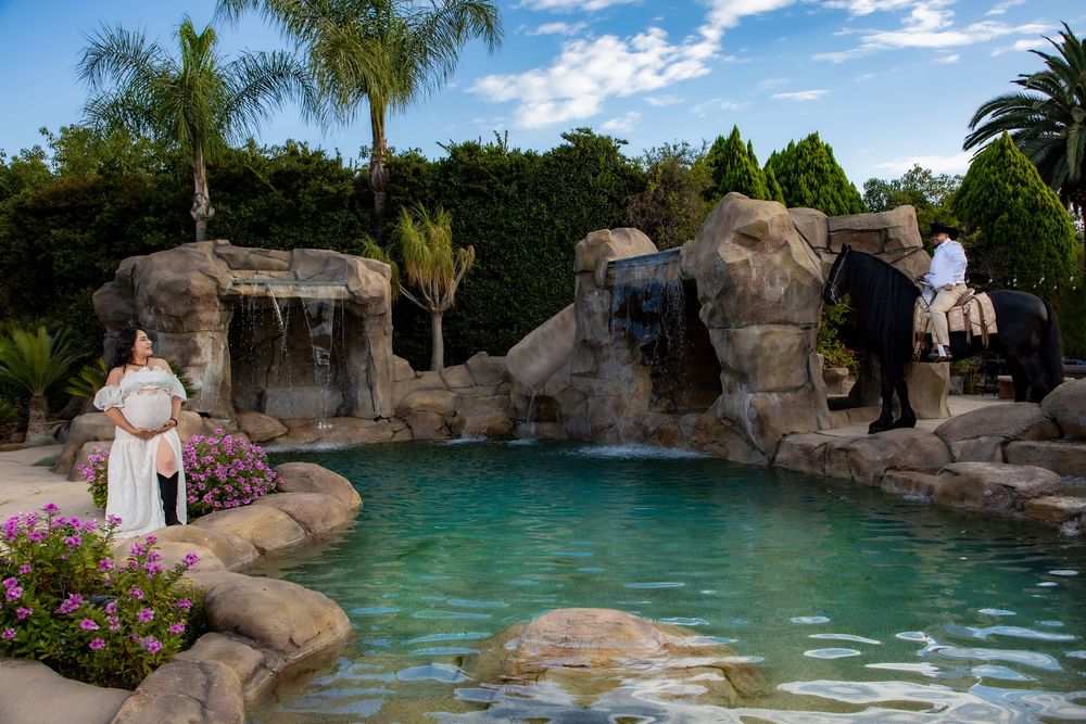 Pregnant woman in white dress stands by a rocky waterfall pool as a man rides a horse.