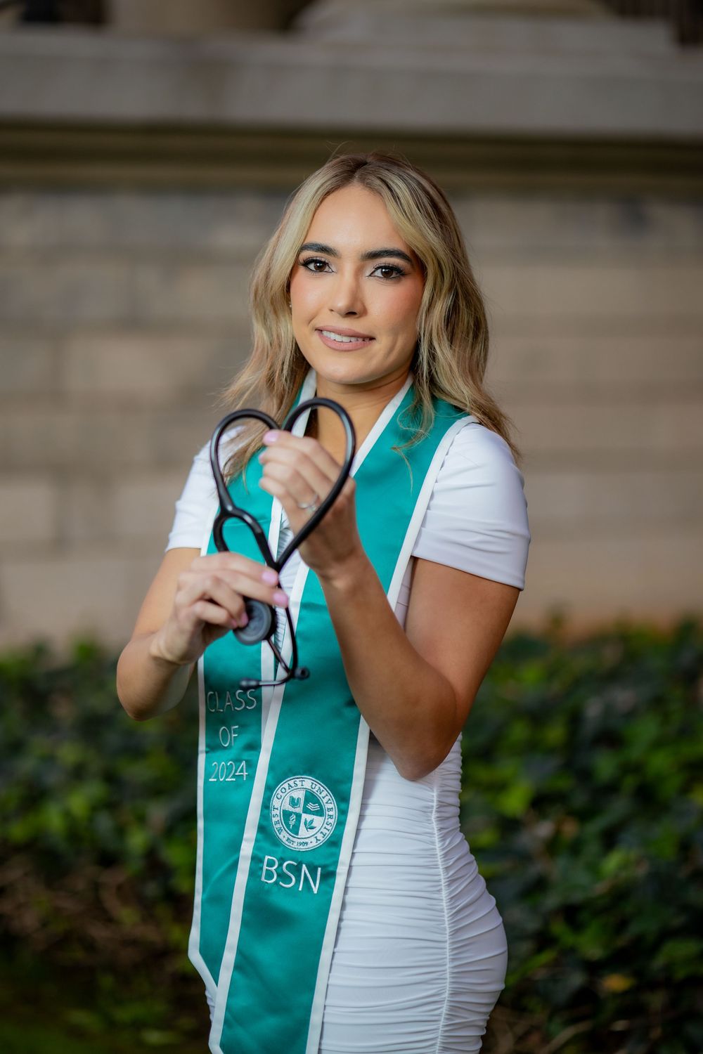 BSN graduate in white dress and teal Class of 2024 stole holds stethoscope forming a heart shape.