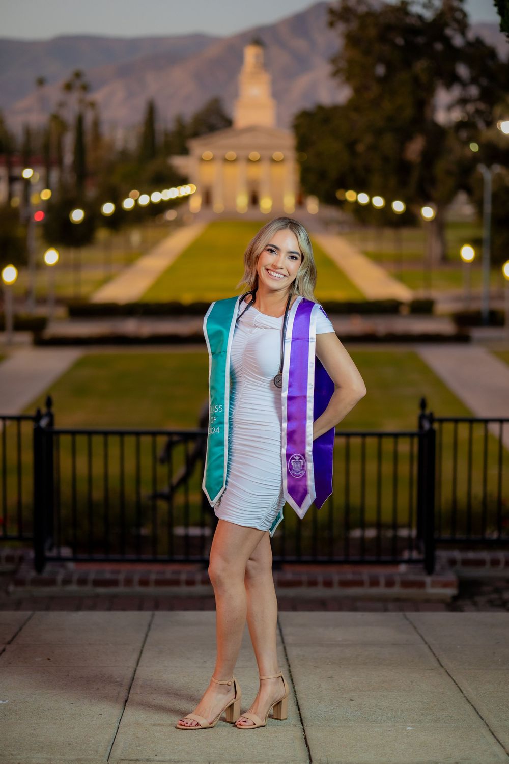 Nursing graduate poses at dusk in white dress with teal BSN and purple stoles before lit campus building.