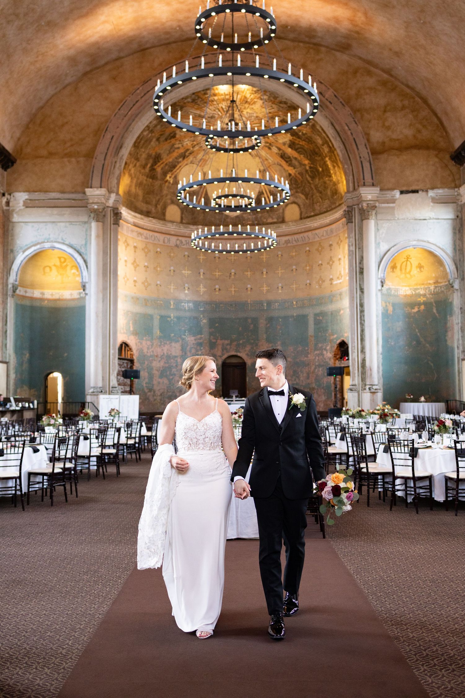 Bride and groom walk hand-in-hand in an elegant ballroom with chandeliers and reception tables set for their wedding.