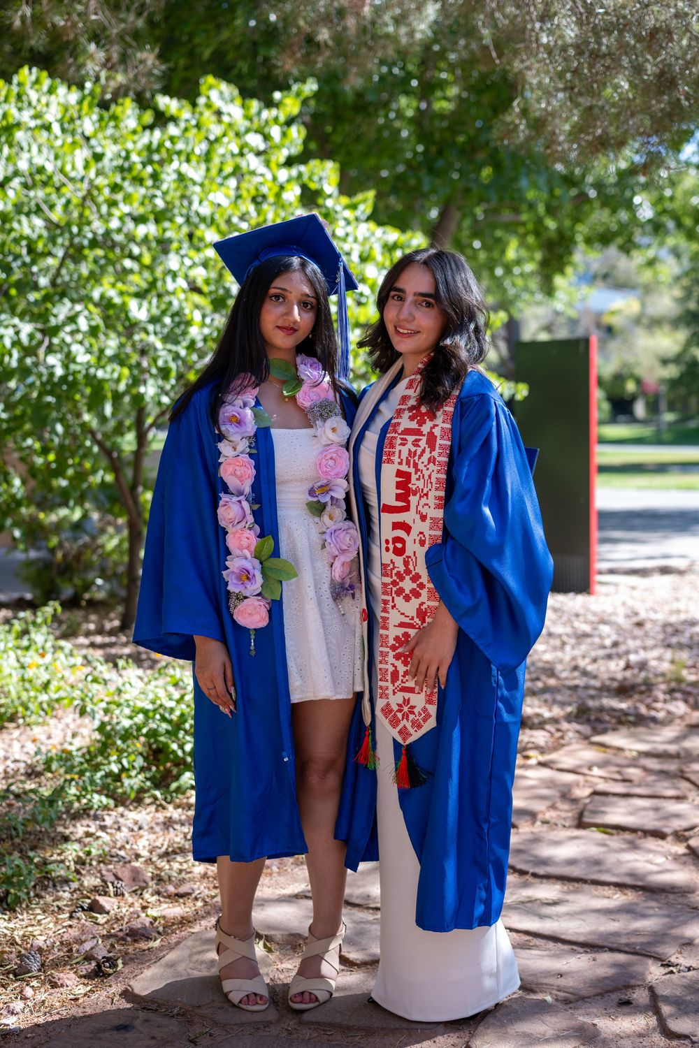 Two female graduates in blue caps and gowns pose outdoors, one wearing a floral lei and the other a red embroidered stole.