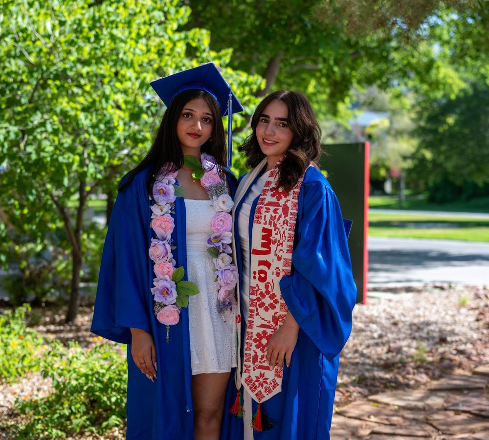 Two female graduates in blue caps and gowns pose outdoors, one wearing floral lei and other a decorative red sash.