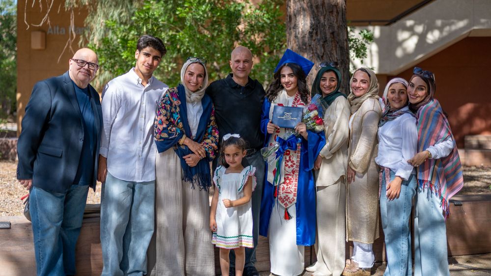 A group of diverse family members posing together outdoors at a graduation celebration with one person in blue cap and gown.
