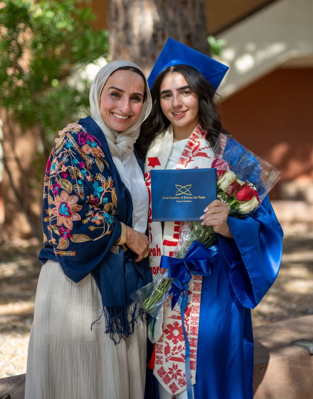 Two women smile at graduation, one in blue cap and gown holding diploma and flowers outdoors.