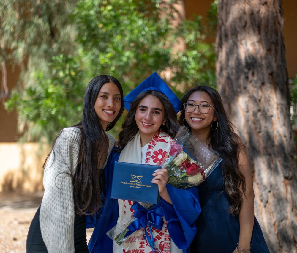 Graduate in blue regalia holds flowers and diploma while posing with two friends outdoors.
