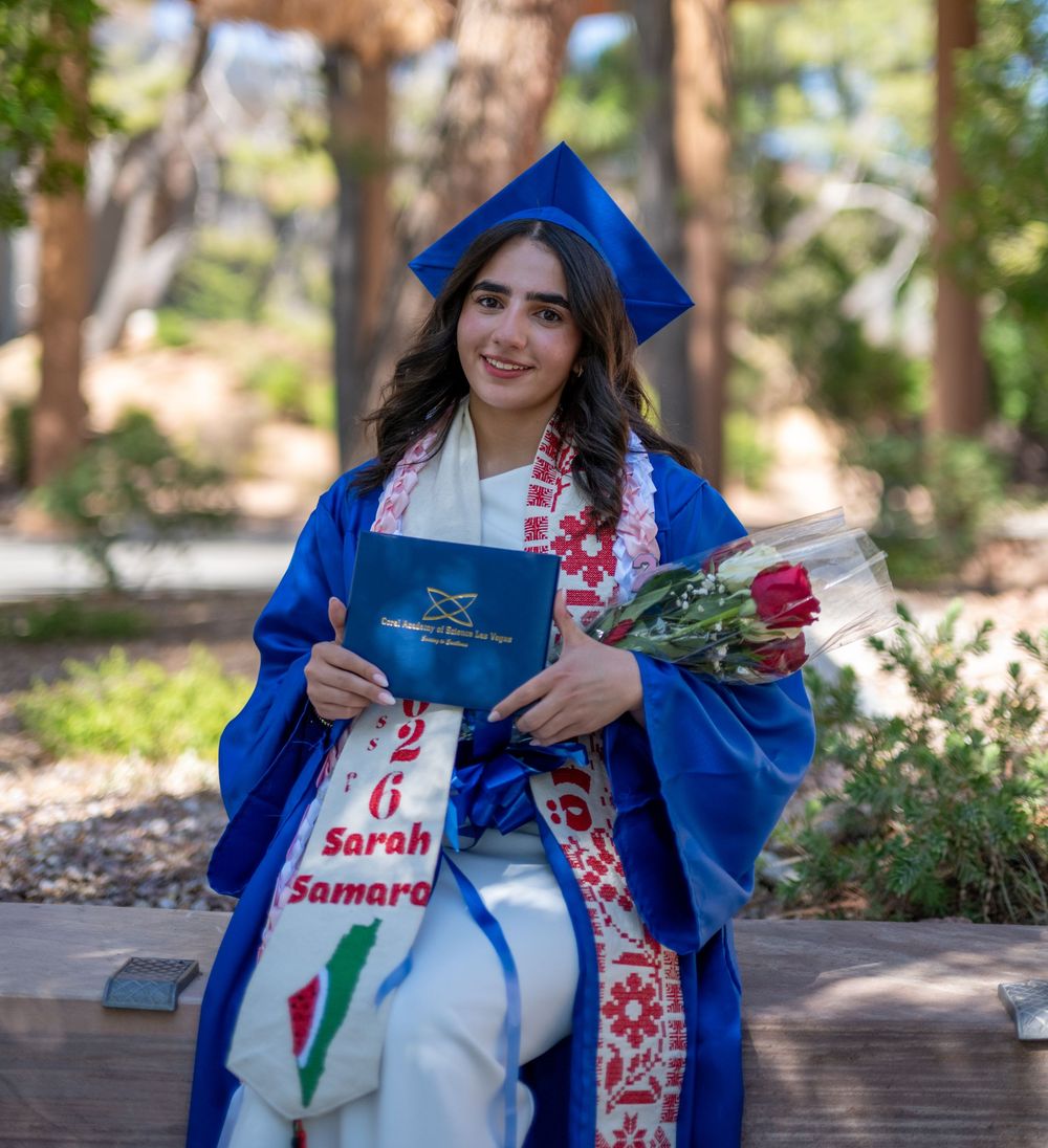 Female graduate in blue cap and gown sits outdoors holding roses with graduation sash reading Sarah Samara.
