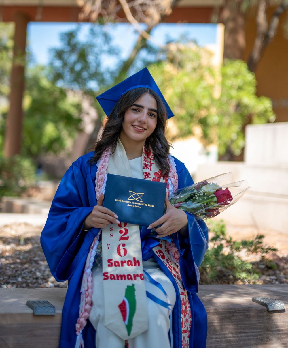 Graduate in blue cap and gown holding diploma and roses, wearing personalized sash with name Sarah Samaro, class of 2026.