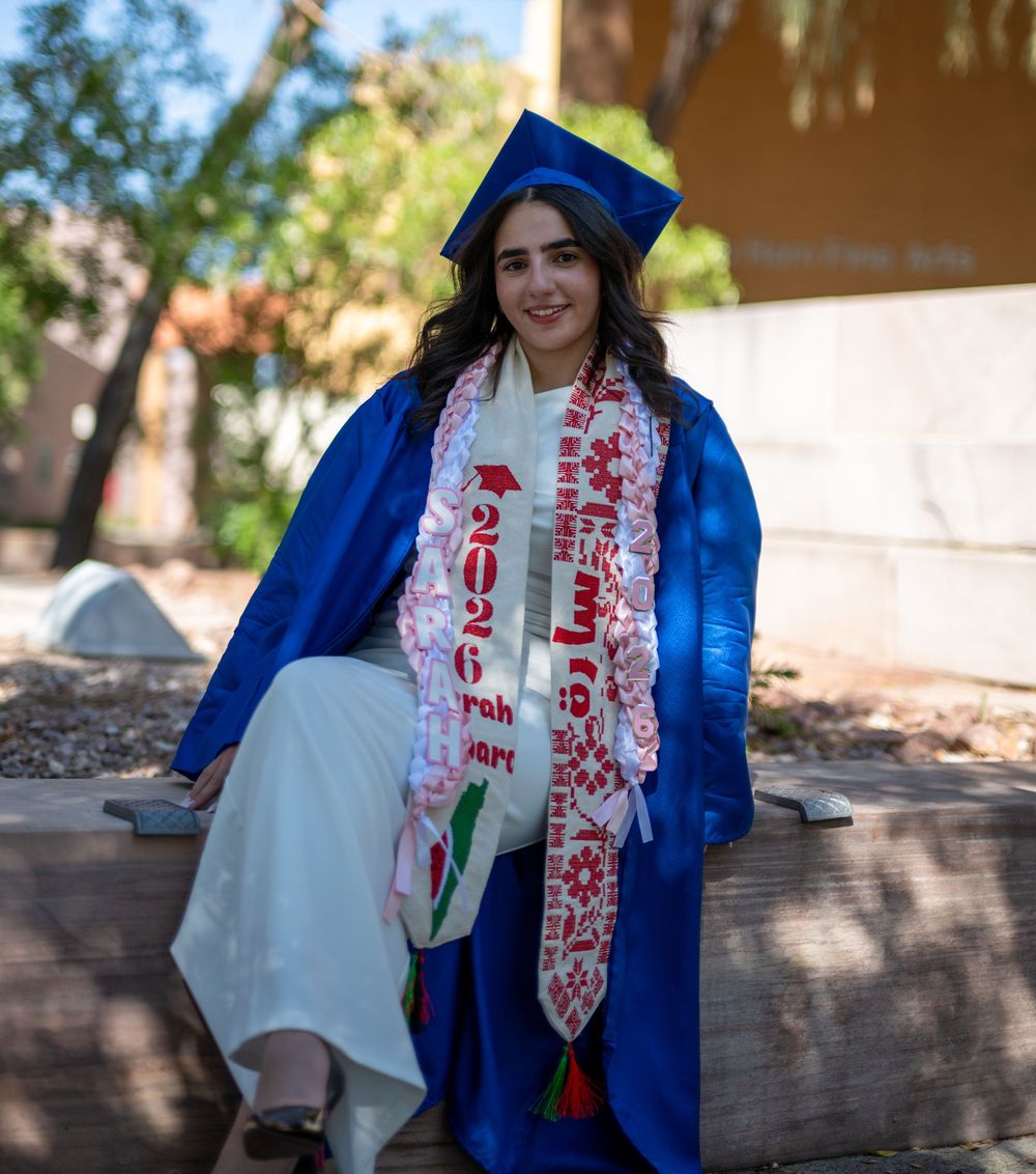 Young woman in blue graduation cap and gown with sash seated outdoors on campus.