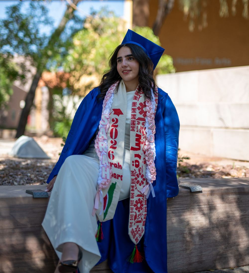 Graduate in blue cap and gown with Class of 2026 sash sits outdoors in sunny setting.