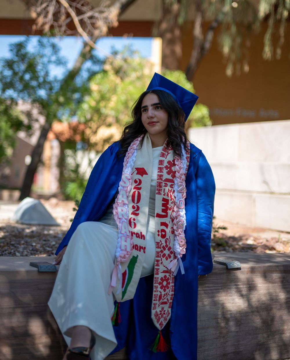 Graduate in blue cap and gown wearing Class of 2026 sash sits on bench in outdoor setting.