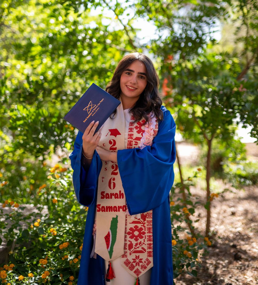 A smiling female graduate in blue gown holds her diploma, wearing a red embroidered Palestinian stole outdoors.