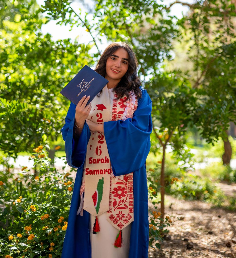 Smiling graduate in blue robe holds diploma among green trees wearing Class of 2026 graduation sash.