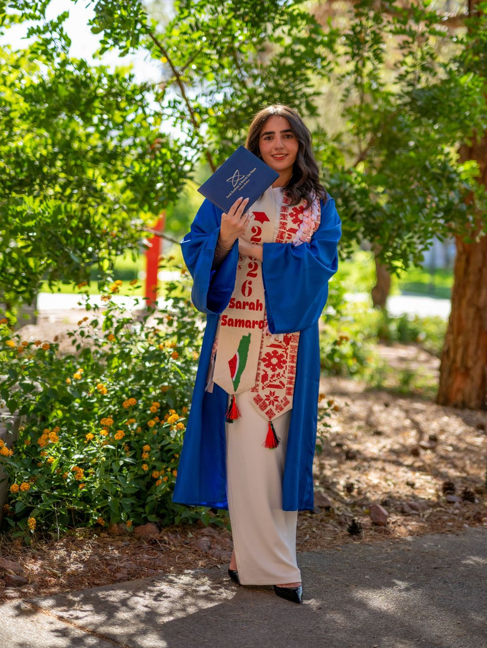 A female graduate in blue gown and embroidered stole poses with diploma on a sunny, garden-lined campus path.
