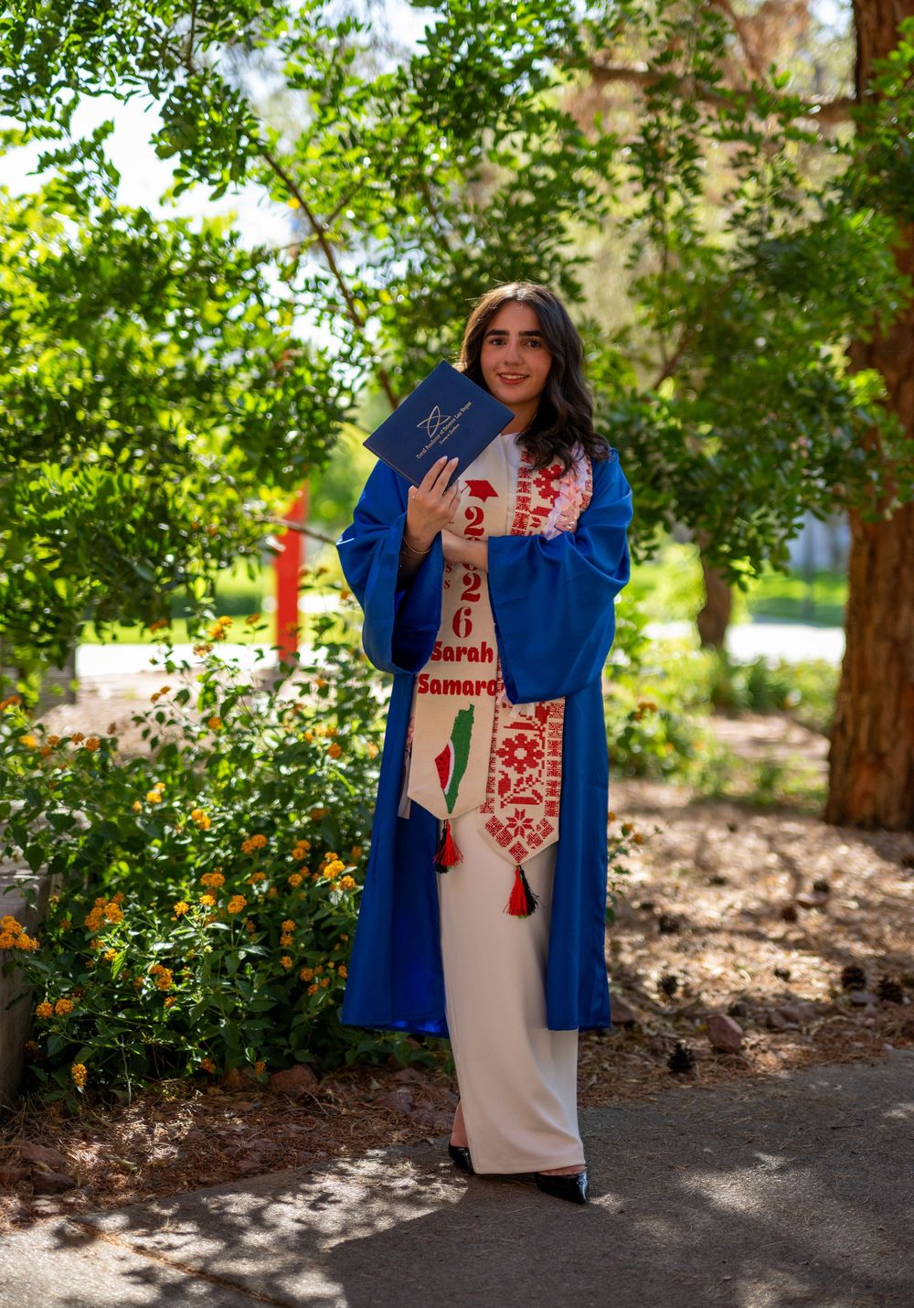 A female graduate in blue commencement gown holds diploma while posing on a tree-lined campus walkway.