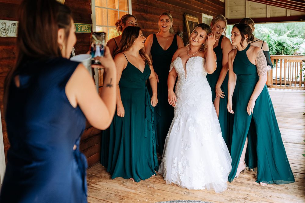 Bride in white lace gown surrounded by bridesmaids in teal dresses inside a rustic wooden barn.