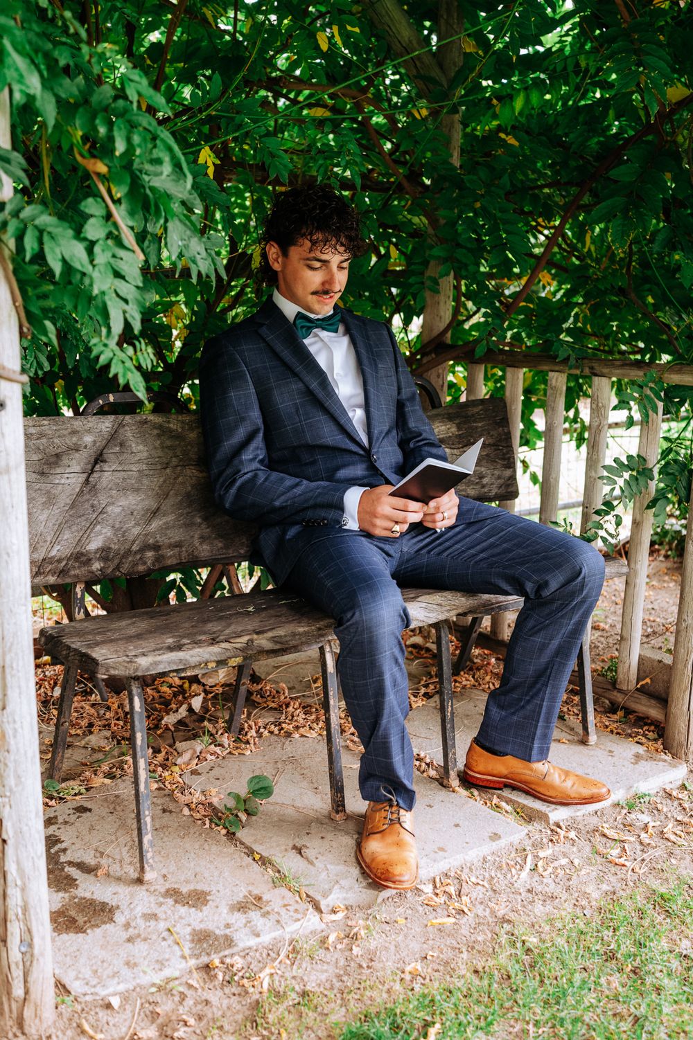 Groom in navy suit with green bow tie reading book on wooden bench under tree outdoors.