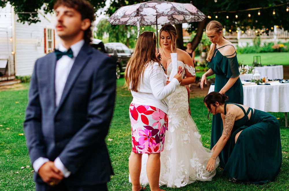 Bride in white lace gown assisted by guests outdoors at elegant garden wedding reception.