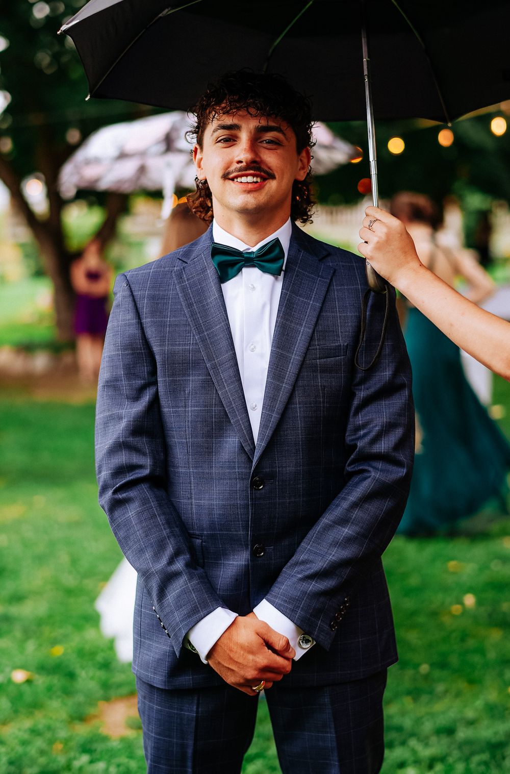 Smiling groom in a navy plaid suit with green bow tie stands under an umbrella at an outdoor wedding.