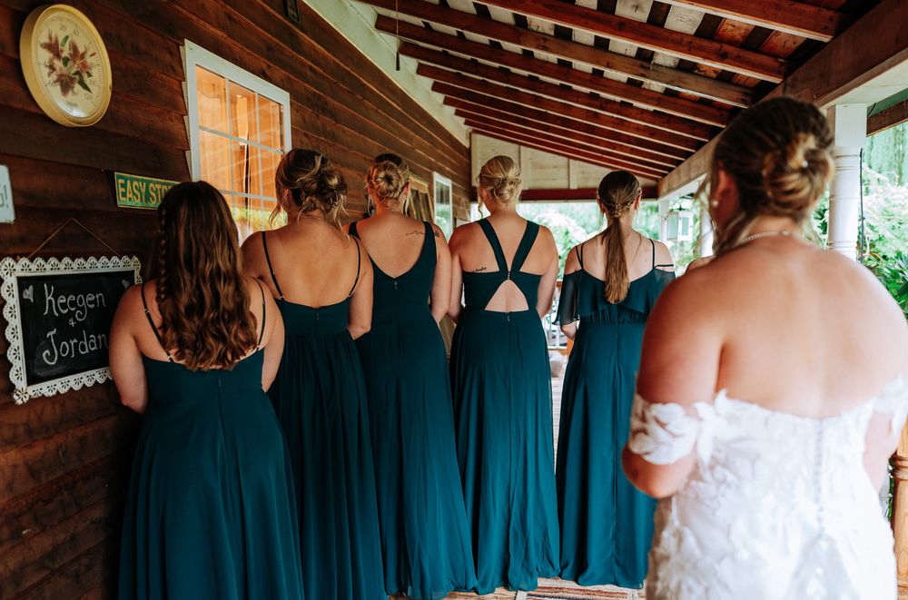 Bridesmaids in teal gowns stand with their backs to the camera beside a bride in white at a rustic wedding.